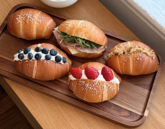 Wooden tray with assorted filled rolls, ham and greens, blueberries, strawberries, and floss—beside a bowl of creamy soup by a window.